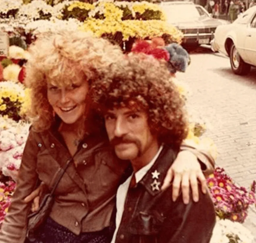 A warm 1970s color snapshot of James McNamara — dark curly hair, mustache, star pins on his jacket — with Lynn Segerblom, her arm around his shoulders, in front of a flower stand.