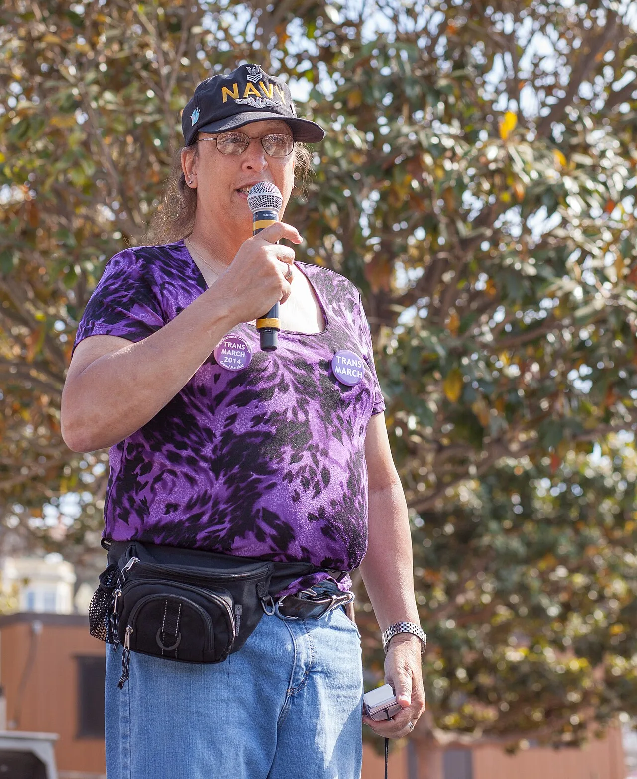Monica Helms speaking into a handheld microphone outdoors, wearing a Navy cap, glasses, and a purple shirt with two Trans March buttons.