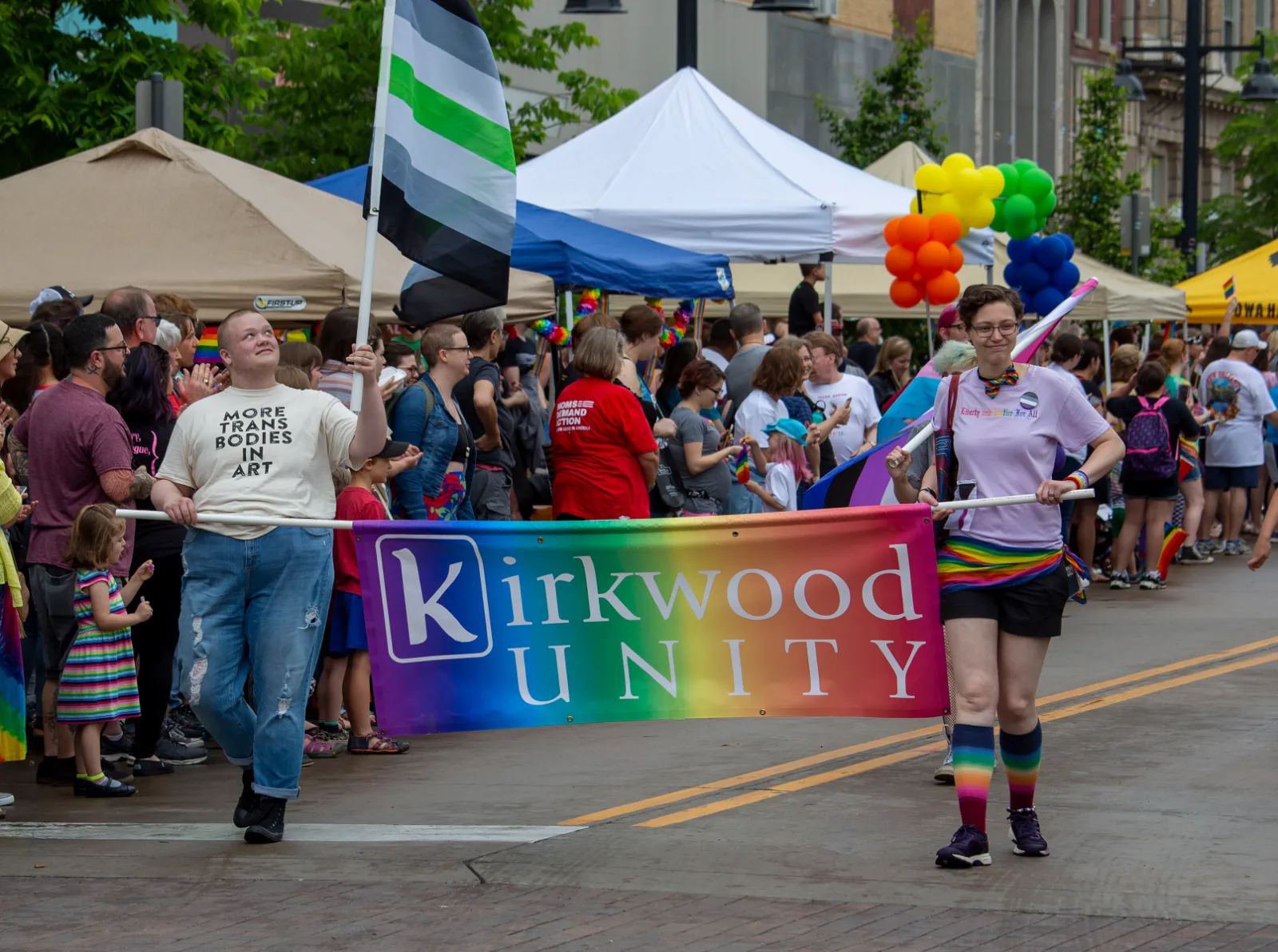 A parade crowd at Iowa City Pride 2019; a marcher carries a large agender flag beside a Kirkwood Unity banner.