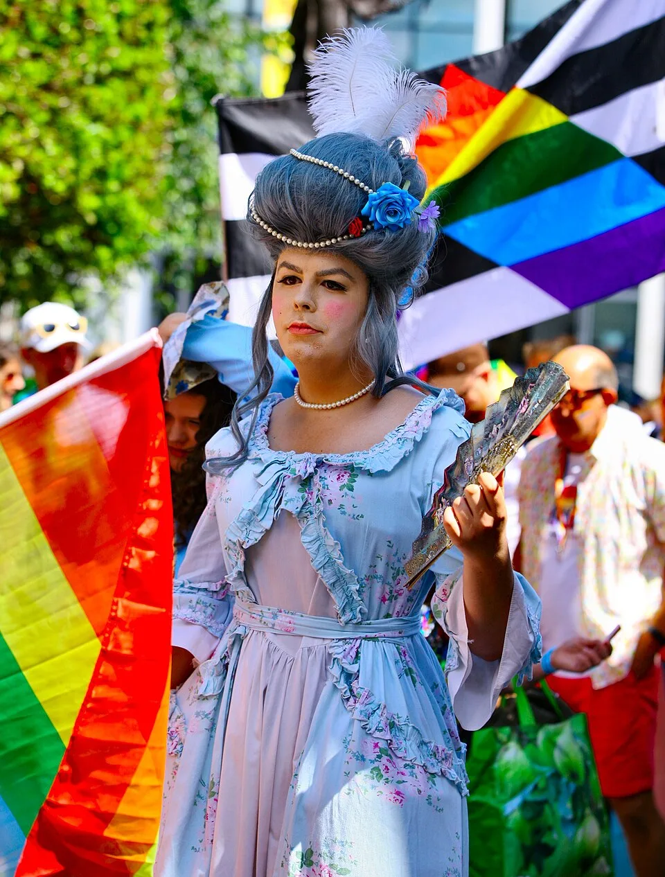 A parade marcher in an eighteenth-century gown of pale blue and pink florals, with a tall grey powdered wig dressed with pearls, white ostrich plumes and a blue rose, holding a painted fan. Behind them a large straight ally flag, black and white stripes charged with a rainbow chevron, is carried beside a rainbow flag.