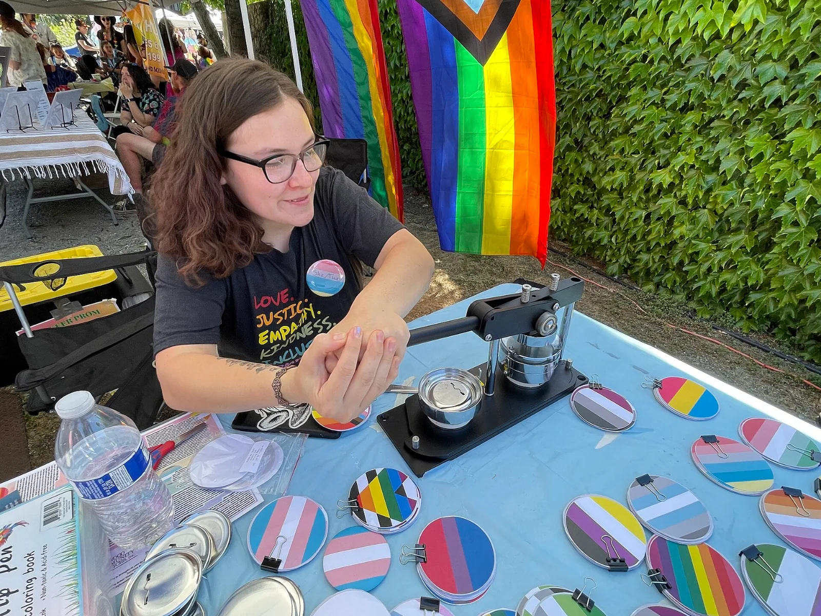 A person in a grey T-shirt printed LOVE. JUSTICE. EMPATHY. KINDNESS. sits at a trestle table under a canopy with a hand-operated button press, wearing a she/they badge. Dozens of finished badges lie on the blue tablecloth in identity flag designs, among them one carrying the straight ally flag's black and white stripes and rainbow chevron. Progress and rainbow flags hang behind.