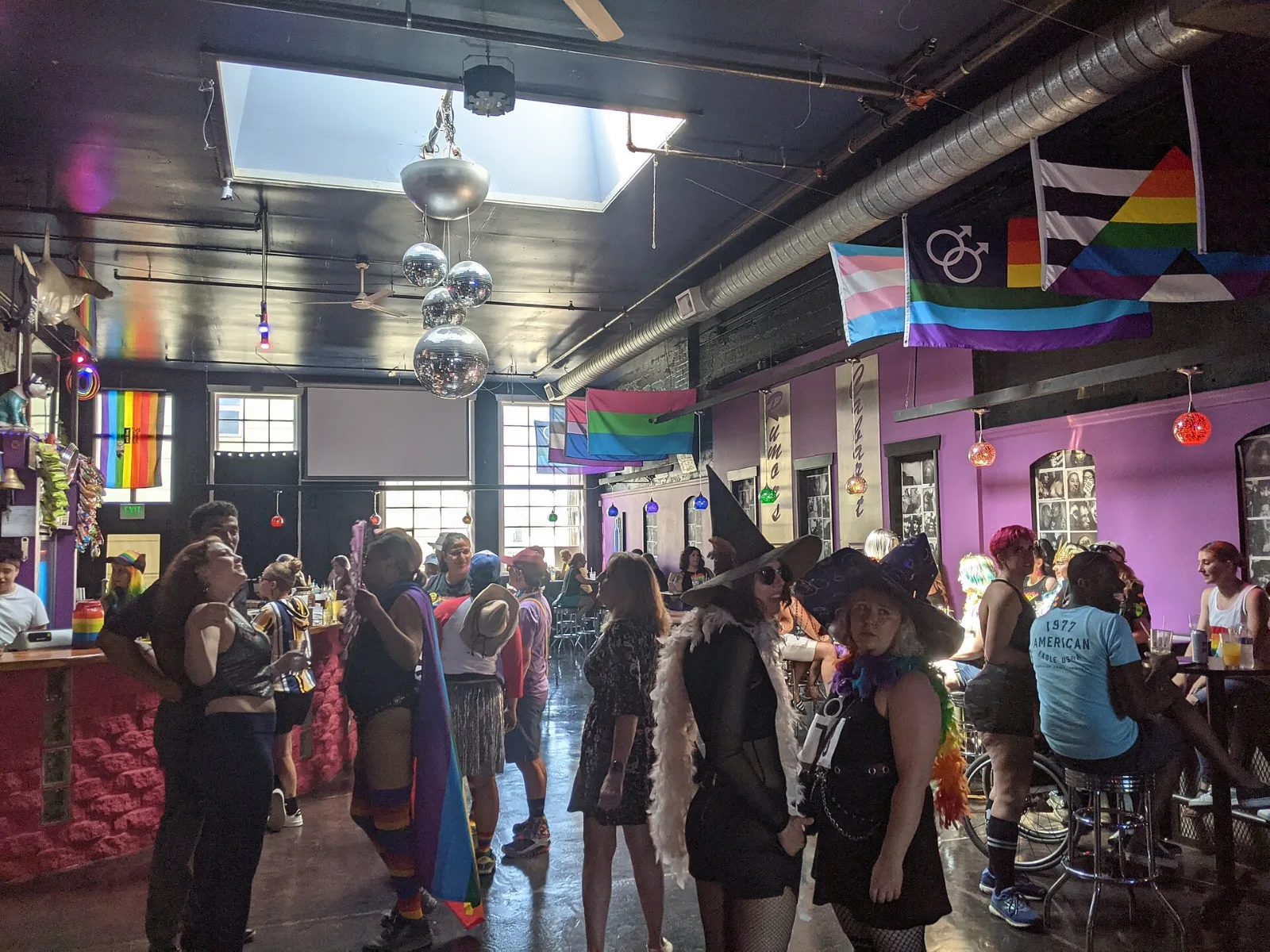 The interior of a bar with purple walls, a skylight and a string of disco balls. Flags hang from a beam near the ceiling: a transgender flag, a dark flag charged with interlocked male symbols, a small orange and pink flag, and at the right the straight ally flag. People stand talking below, several in costume, two in pointed witches' hats.