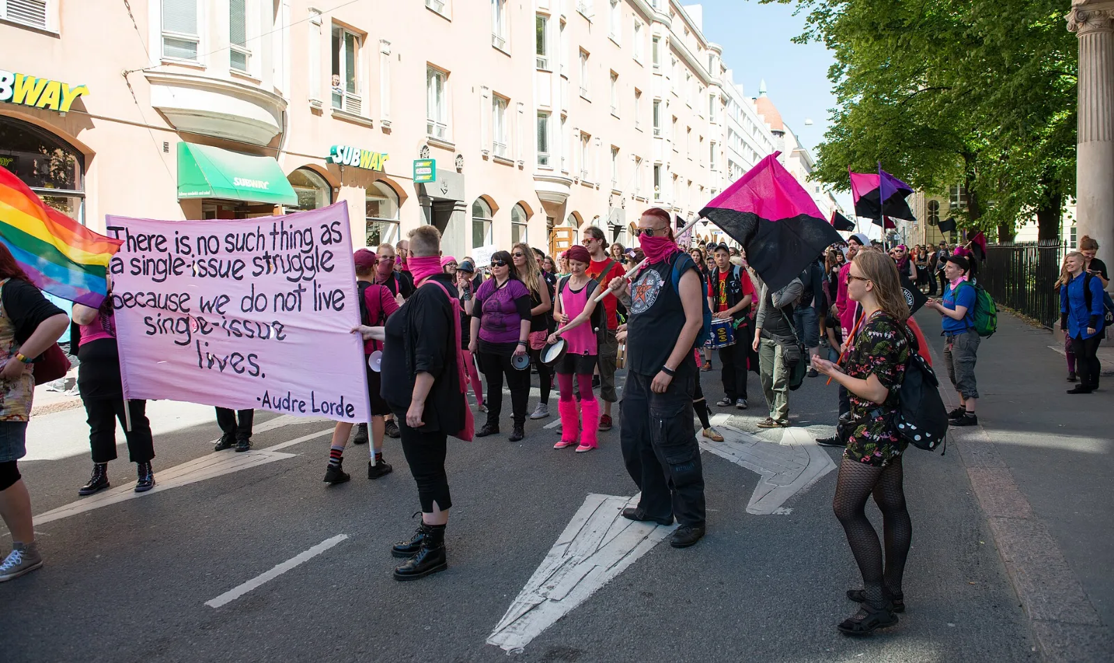 Helsinki Pride marchers carrying a pink and black diagonal flag beside a banner.