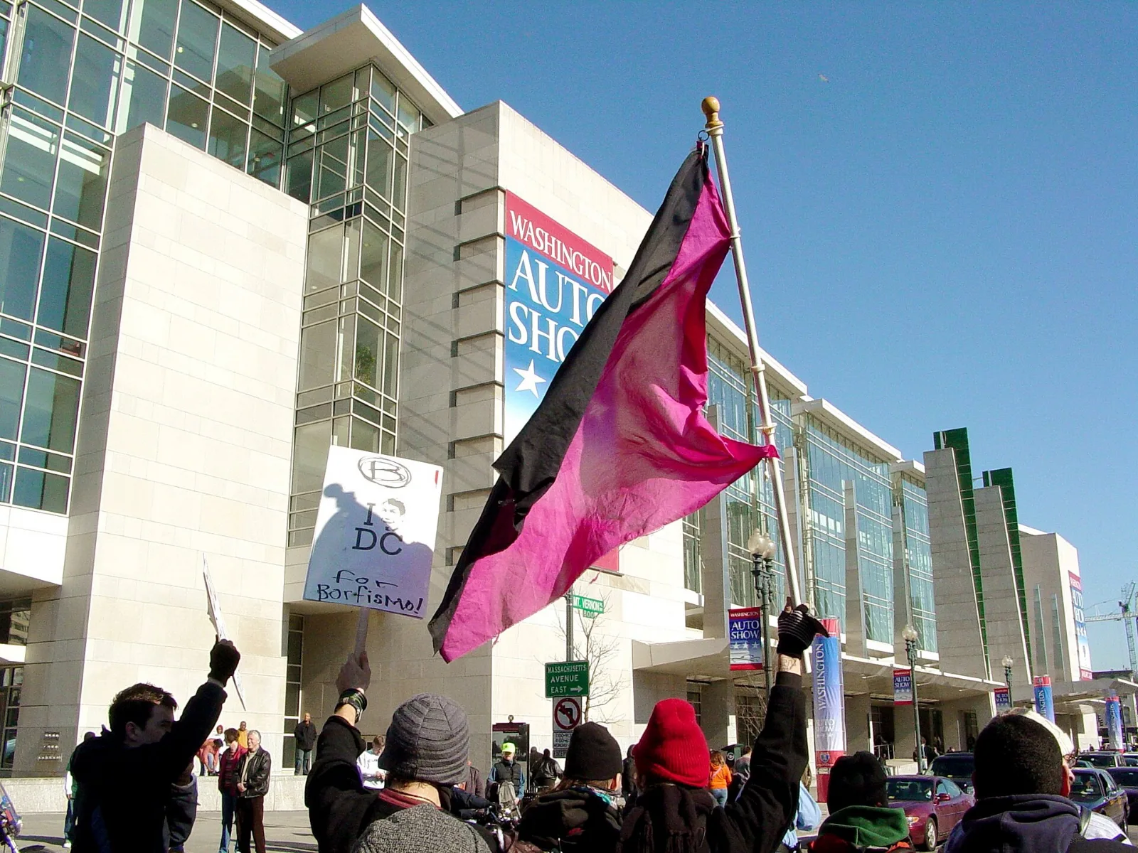 A feeder march passing the Washington Convention Center, a black and pink diagonal flag among the banners.
