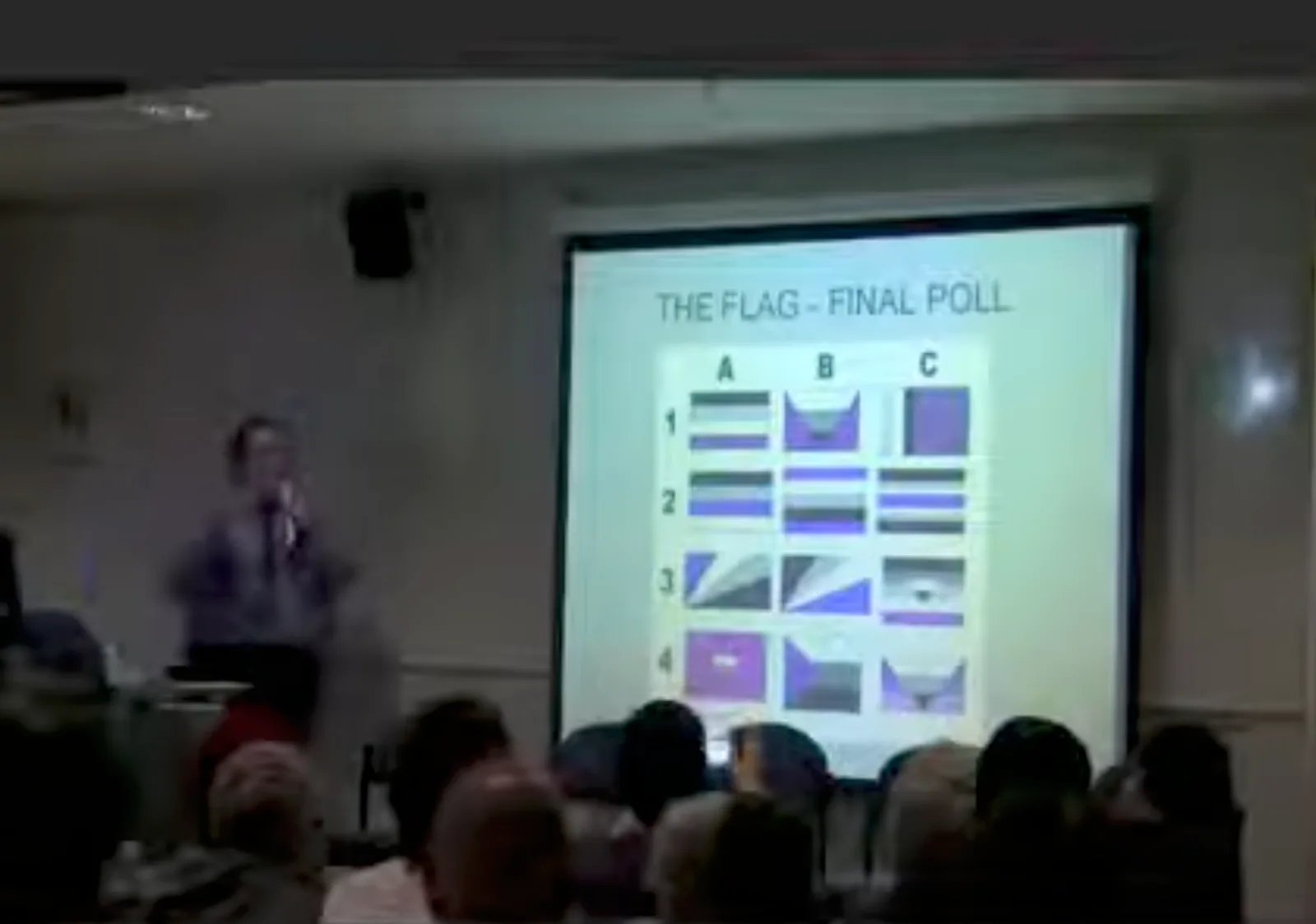 A projection screen in a lecture room showing a slide headed "THE FLAG – FINAL POLL", filled with a grid of small flag designs in black, grey, white and purple, with the speaker beside it.