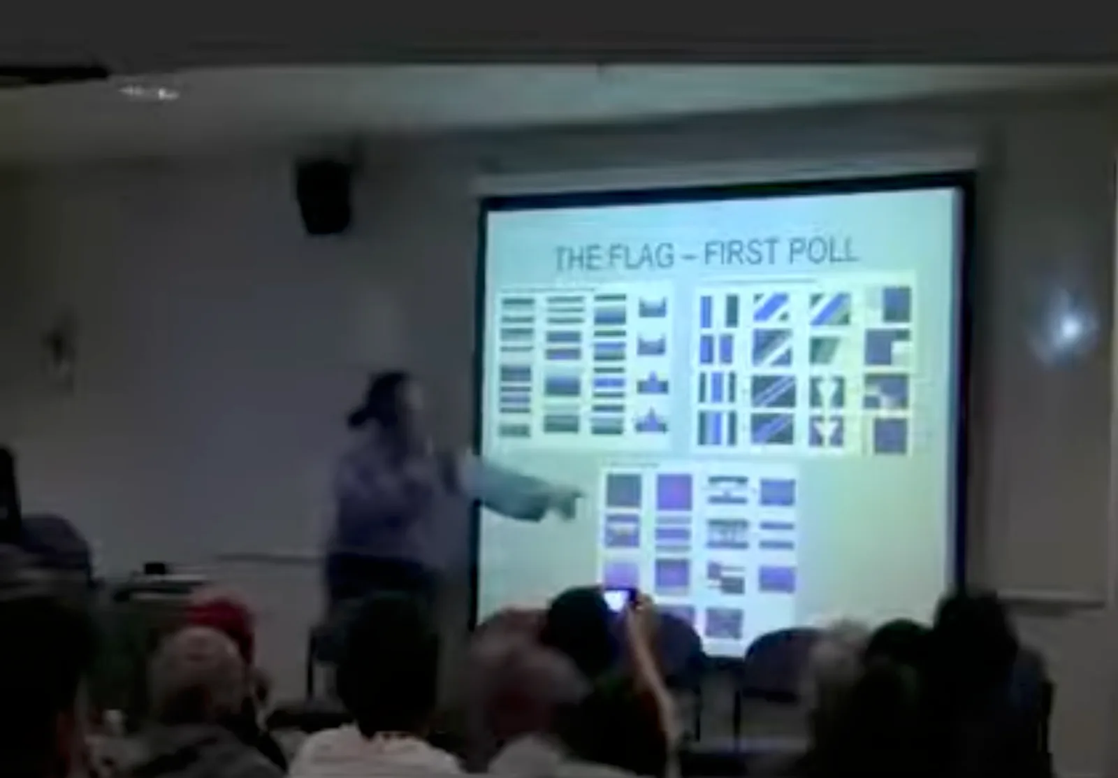 A projection screen in a lecture room showing a slide headed "THE FLAG – FIRST POLL", filled with a grid of small flag designs in black, grey, white and purple, with the speaker beside it.