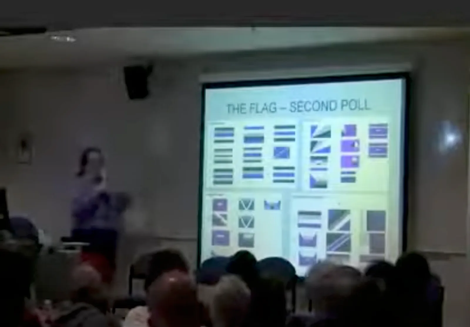 A projection screen in a lecture room showing a slide headed "THE FLAG – SECOND POLL", filled with a grid of small flag designs in black, grey, white and purple, with the speaker beside it.