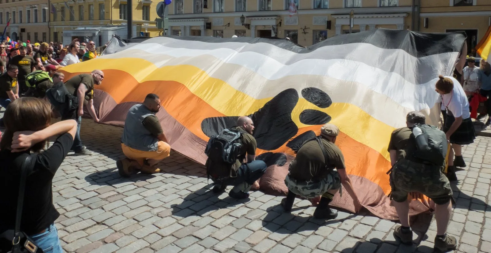 An oversized bear flag carried flat above the heads of marchers across a cobbled square.