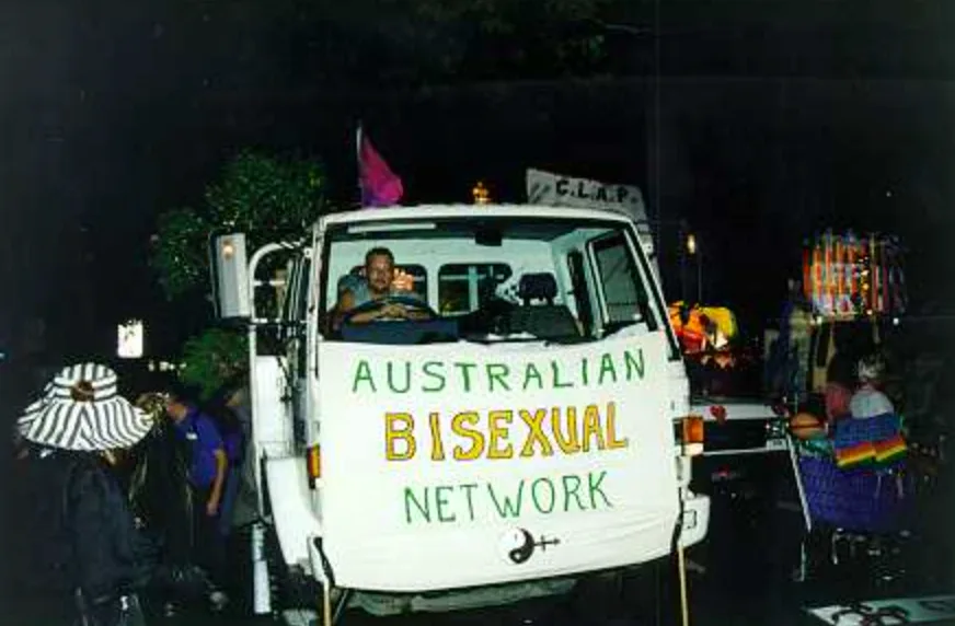 A night photograph of a small white truck with a driver leaning from the cab, its front draped with a hand-painted banner reading "AUSTRALIAN BISEXUAL NETWORK" in green and yellow with a combined gender symbol below.