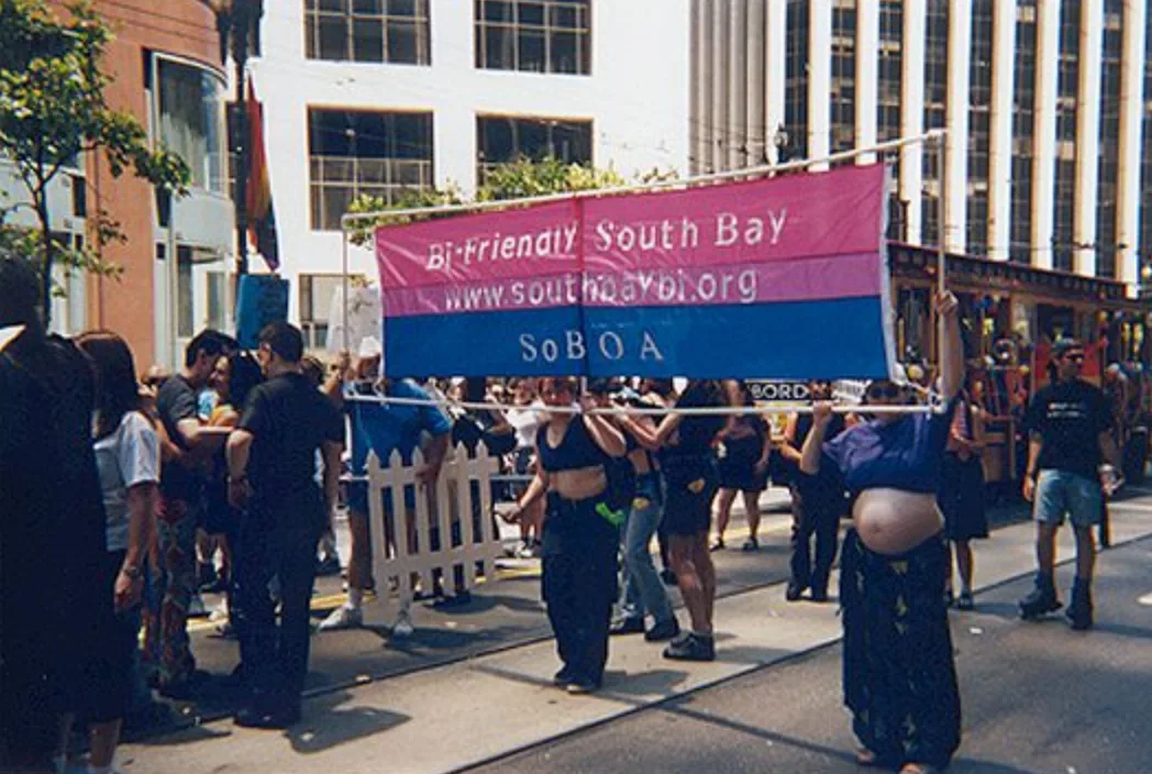 Marchers on a city street carrying a wide banner in magenta, lavender and blue reading "Bi-Friendly South Bay / www.southbaybi.org / SoBOA", with a streetcar behind them.