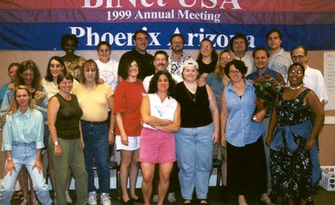 About twenty people posed in rows beneath a large banner in magenta, lavender and blue reading "BiNet USA 1999 Annual Meeting, Phoenix Arizona".