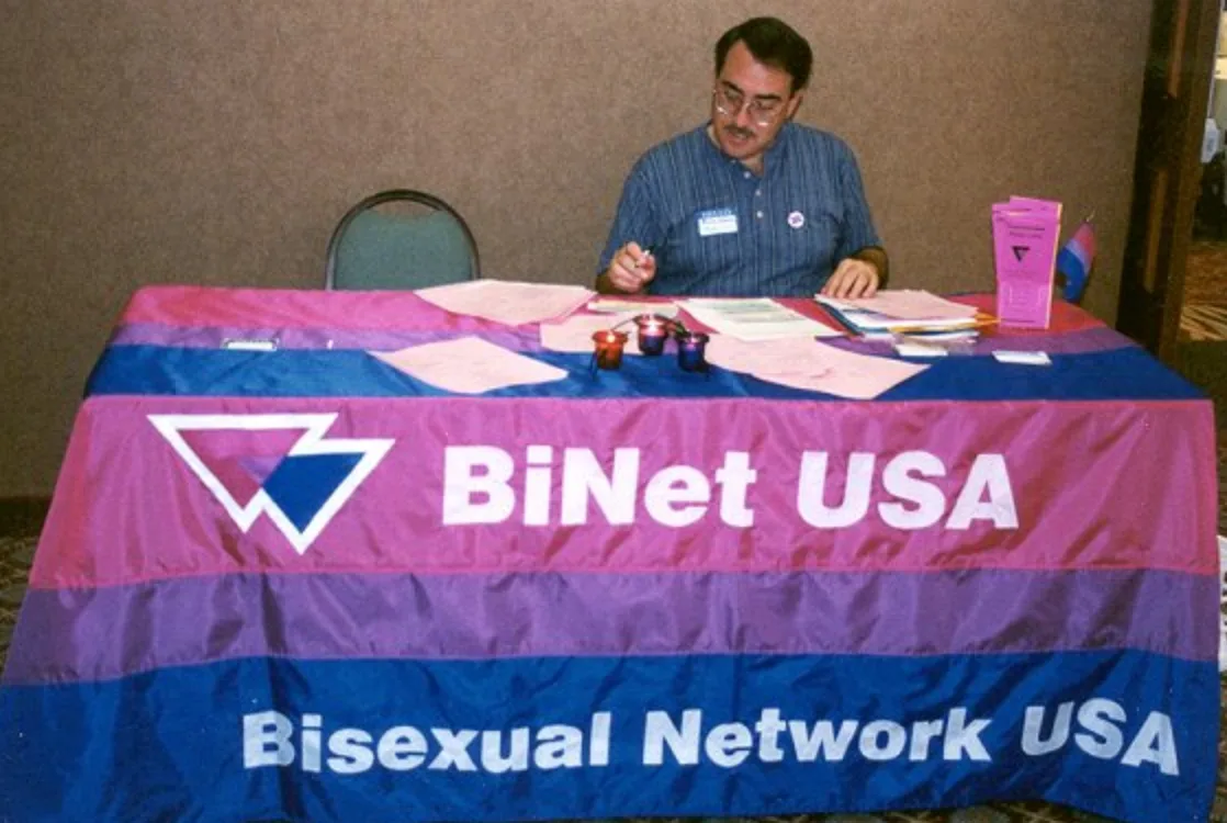 A man in a blue striped shirt and glasses sits writing at a trestle table draped in a magenta, lavender and blue cloth reading "BiNet USA / Bisexual Network USA", with a small bisexual flag on a stand beside him.