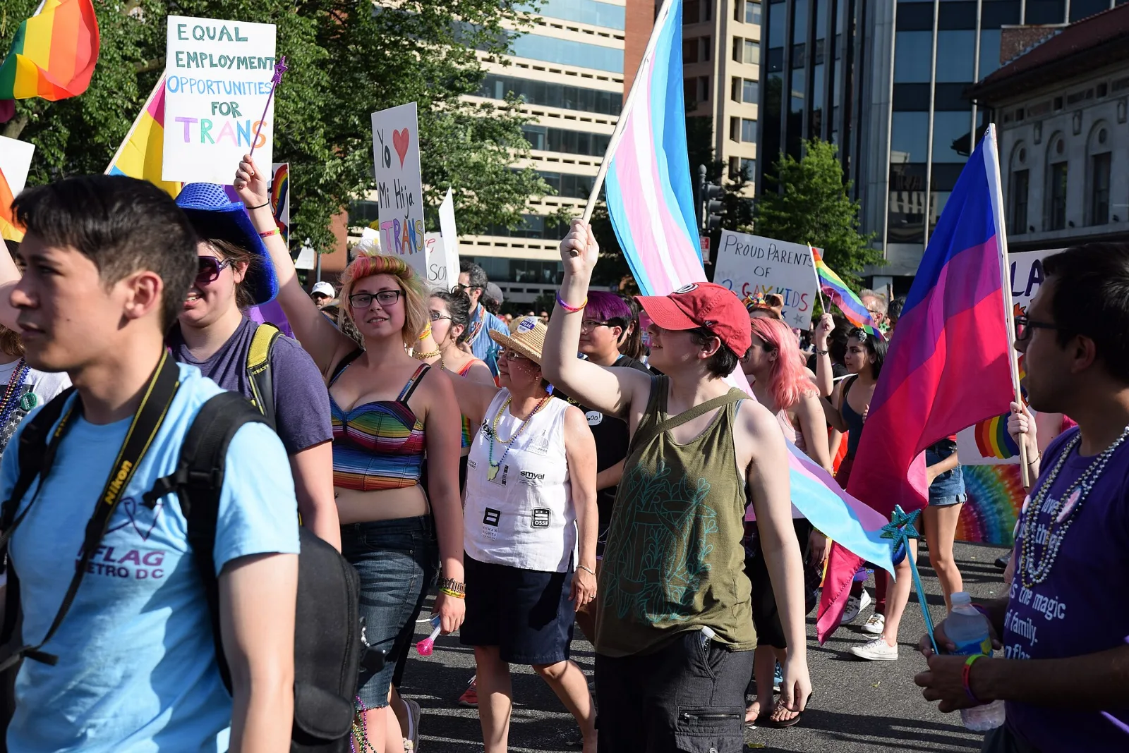 A crowded Pride parade with a large bisexual flag at right, a large transgender flag at center, rainbow and pansexual flags, and hand-lettered placards about trans rights.
