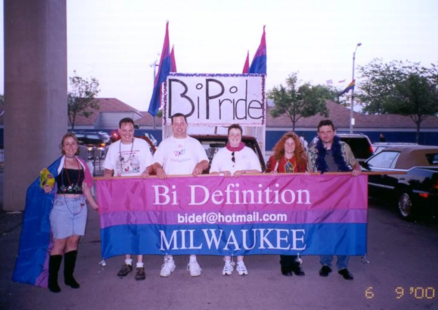 Six people behind a banner in magenta, lavender and blue reading "Bi Definition / bidef@hotmail.com / MILWAUKEE", with three bisexual flags on poles and a hand-lettered BiPride float sign behind them.