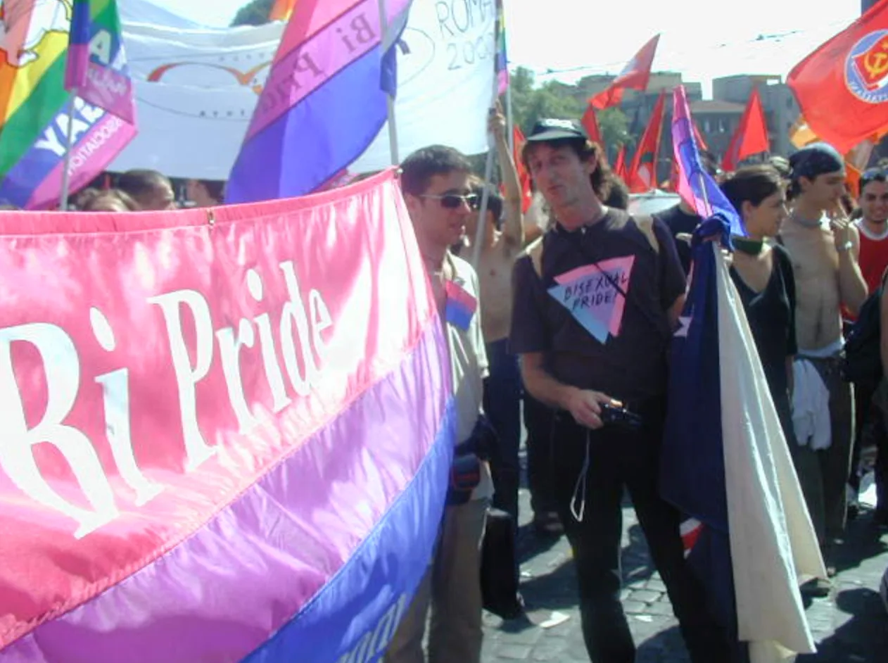 A close view of the "Bi Pride" banner in the foreground, with a man in a black cap and a t-shirt printed with a pink and blue "Bisexual Pride" triangle standing beside it holding a camera, among red flags and a "ROMA 2000" banner.