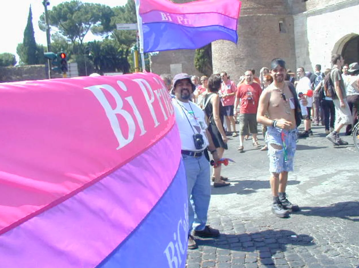The furled Bi Pride banner in the foreground with a bisexual flag flying above it, marchers gathered beside a Roman brick gate and tower.