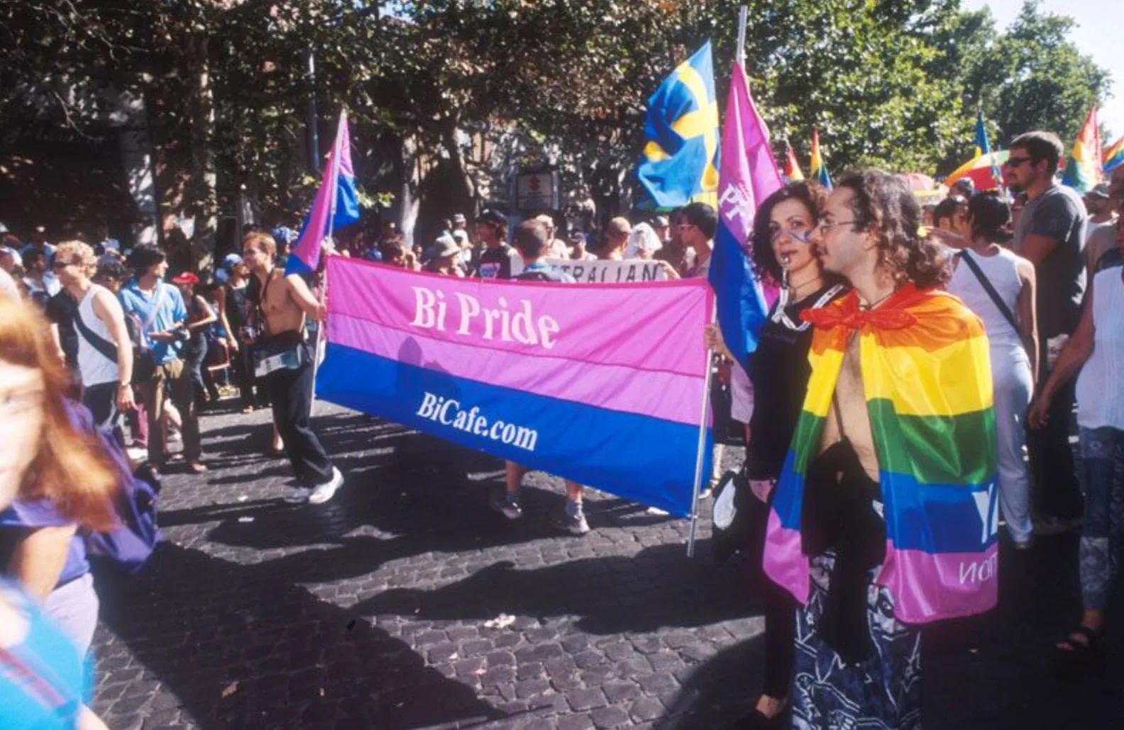 Marchers on a cobbled Roman street carrying a wide banner in magenta, lavender and blue reading "Bi Pride / BiCafe.com", with bisexual flags, a Swedish flag and a rainbow-draped couple alongside.