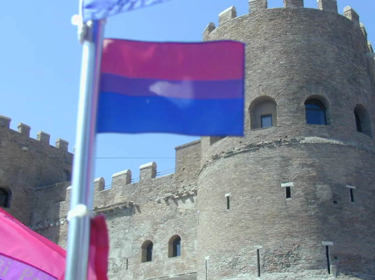 A bisexual pride flag on a metal pole flying against a crenellated brick tower and curtain wall under a clear sky.