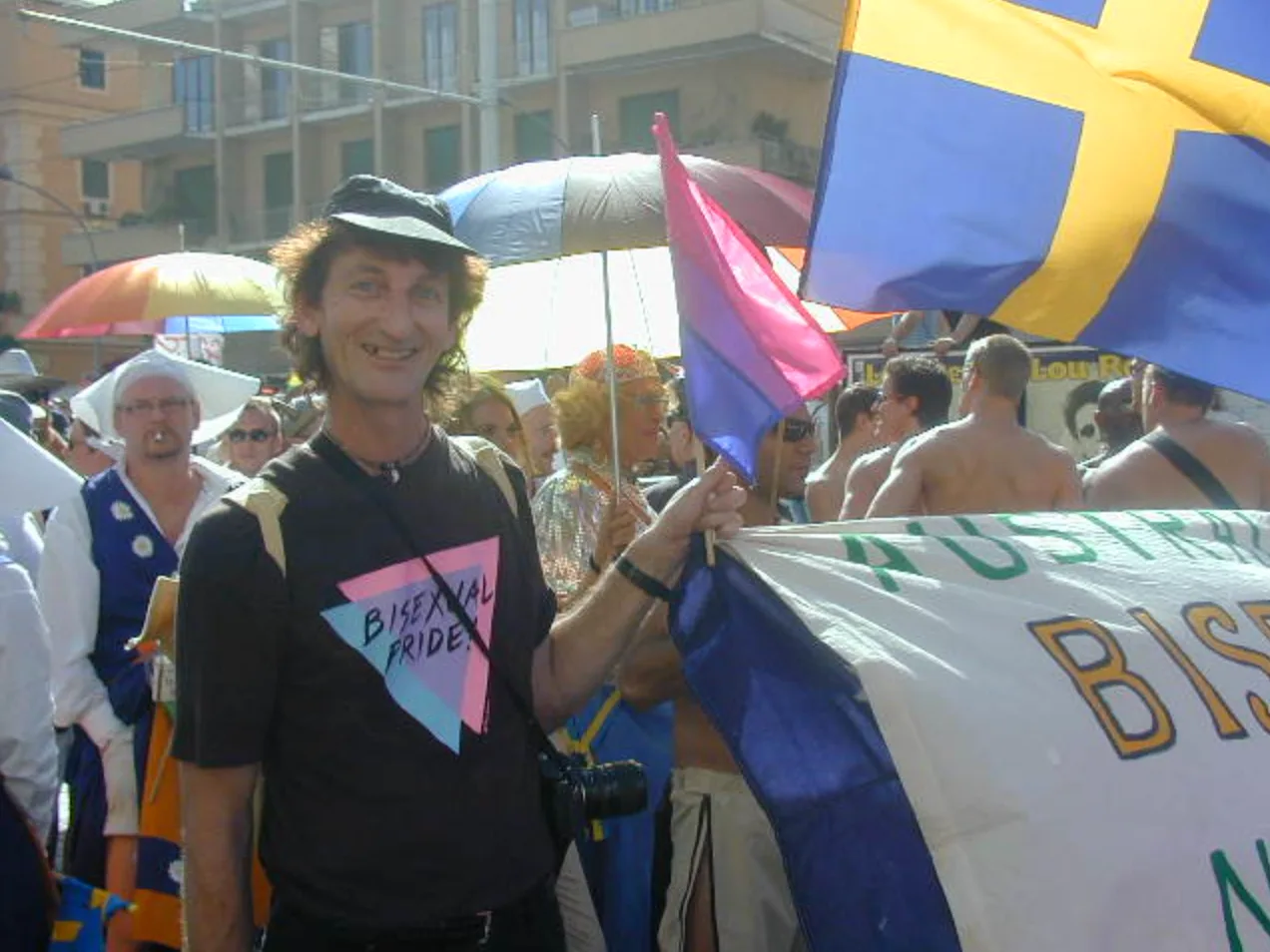 A smiling man in a black cap and a "Bisexual Pride" triangle t-shirt holds a small bisexual flag beside a large hand-lettered white banner reading "AUSTRAL… BISE…", with a Swedish flag above and a camera slung at his hip.