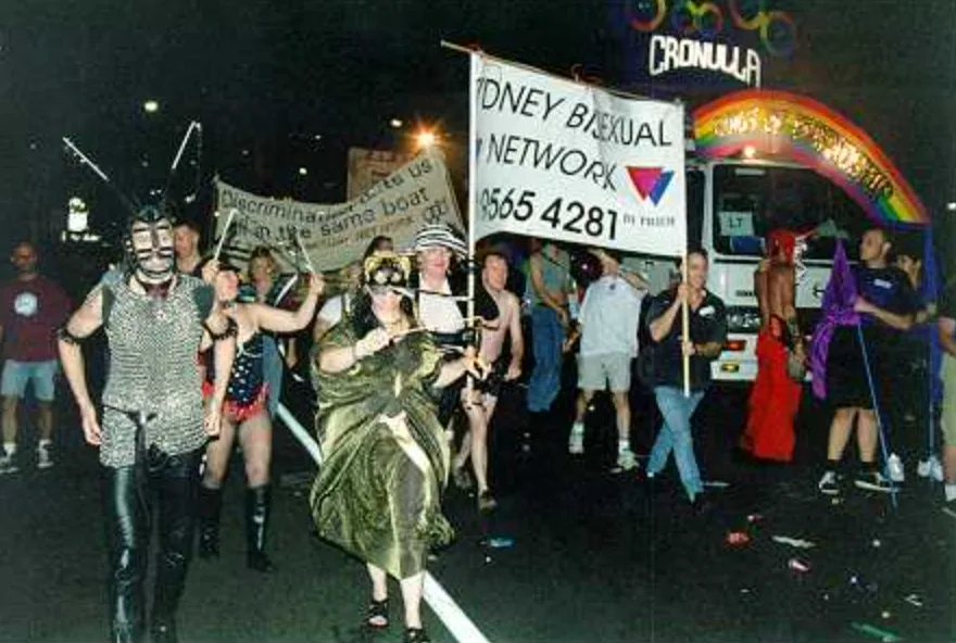 A night parade with costumed marchers, behind them a white banner reading "SYDNEY BISEXUAL NETWORK / 9565 4281 / BI PRIDE" carrying an overlapping pink and blue triangle mark.