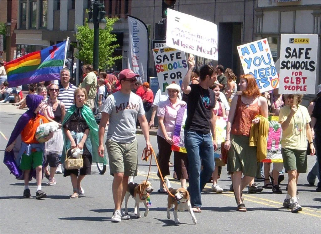 A rainbow flag with a single-blue canton and red cross carried at a Pride march in Portland, Maine, beside marchers holding GLSEN placards reading SAFE SCHOOLS FOR GLBT PEOPLE.