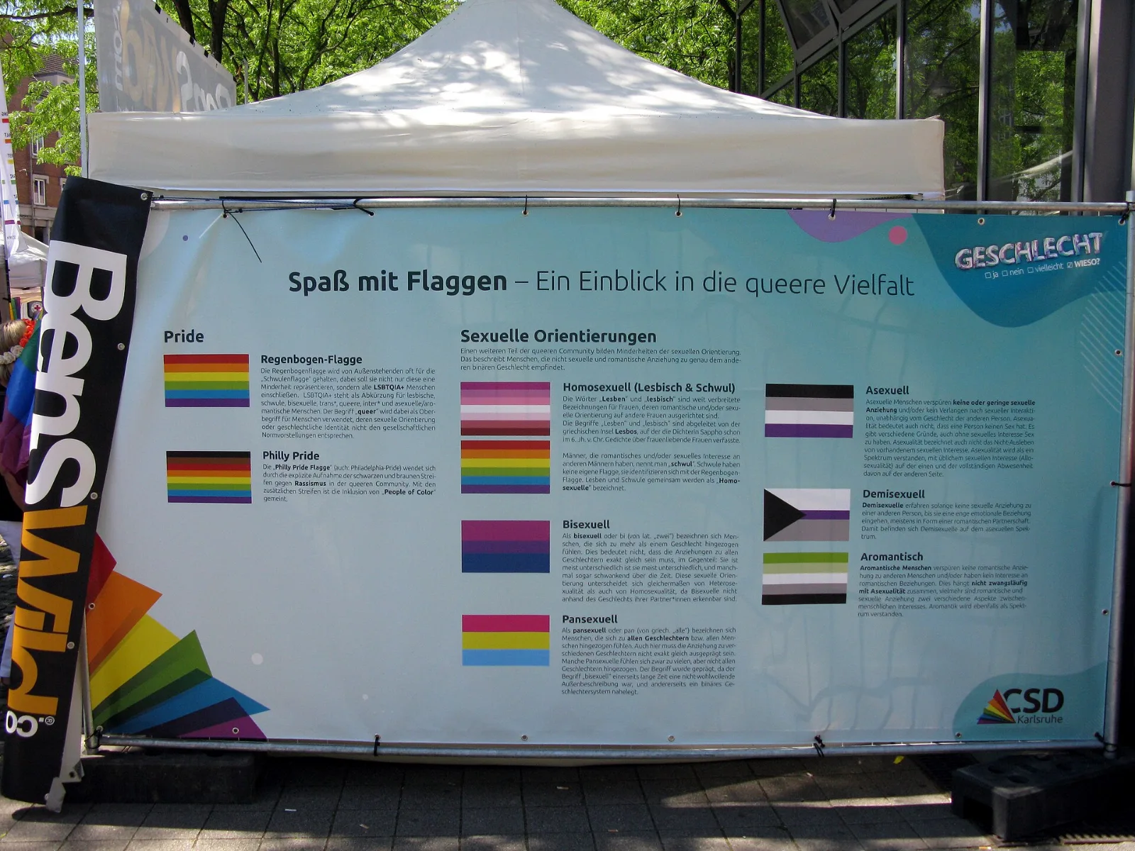 A large printed banner at an outdoor Pride stall headed "Spaß mit Flaggen – Ein Einblick in die queere Vielfalt", showing the rainbow, Philadelphia, lesbian, bisexual, pansexual, asexual, demisexual and aromantic flags each with a paragraph of German explanation.