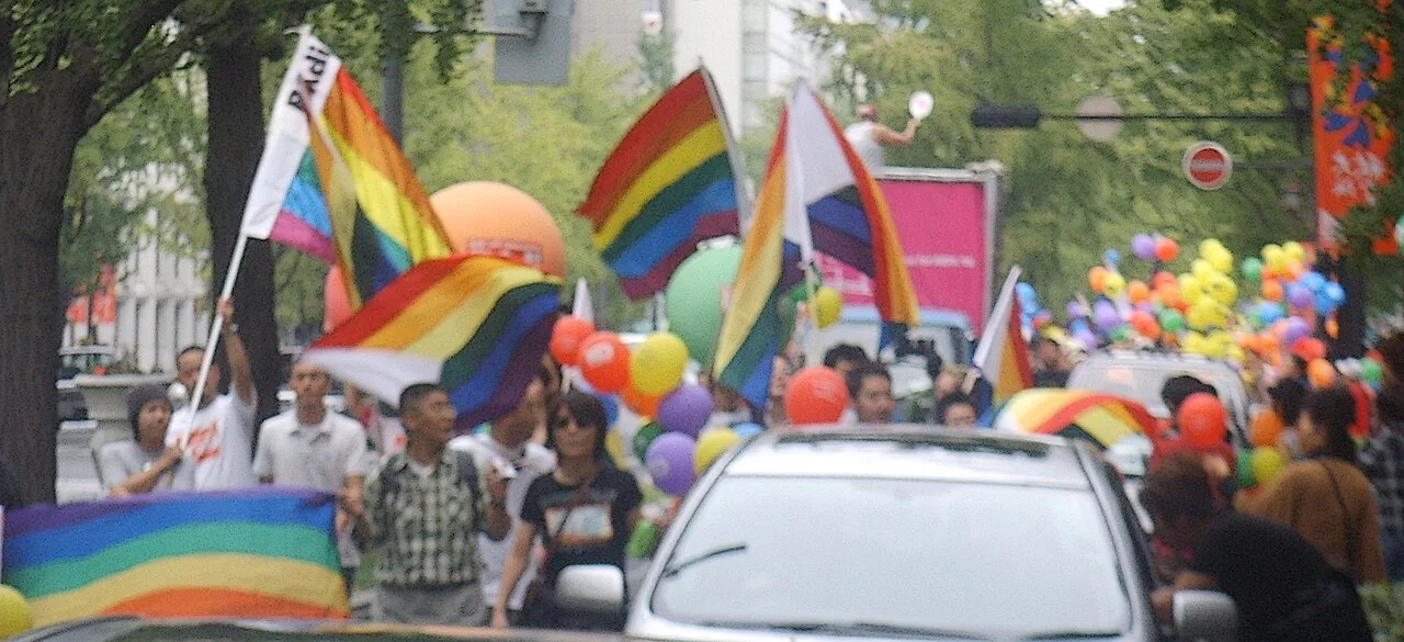 Marchers on a tree-lined Osaka avenue among balloons and a parade truck, carrying rainbow flags, several of which have a white triangle at the hoist.