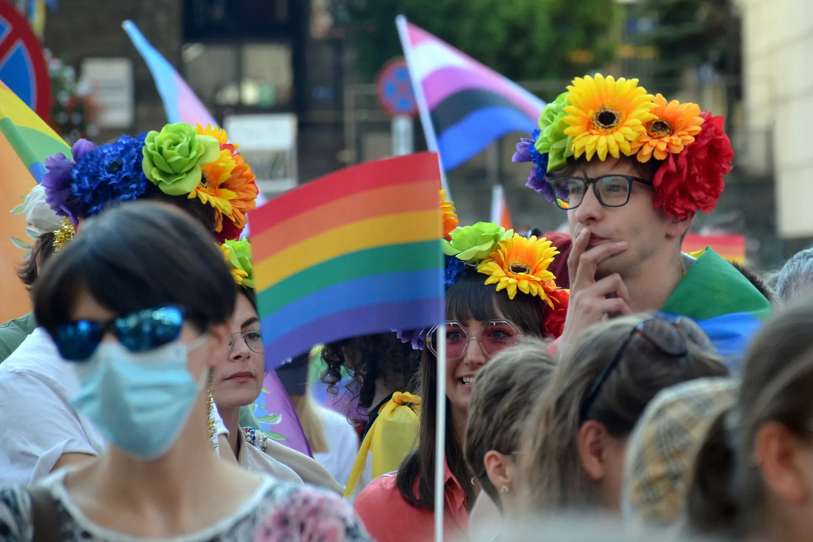 A crowd at a march, several people wearing flower crowns. Behind them a five-stripe flag in pink, white, purple, black and blue is held up beside a rainbow flag.