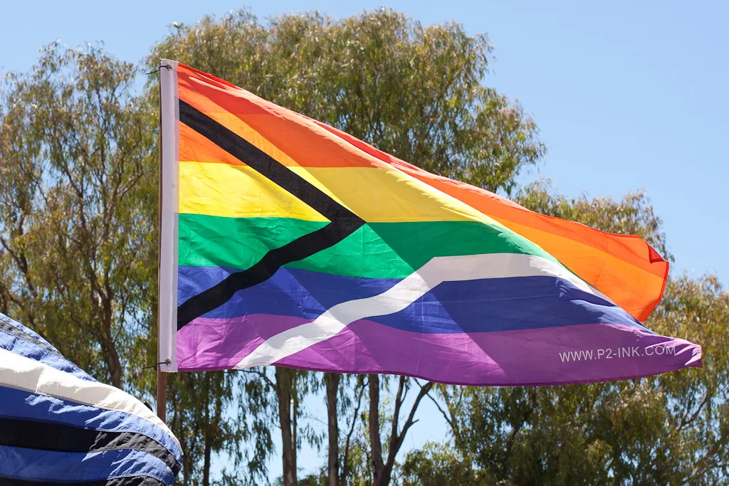 A manufactured flag on a pole against a blue sky and gum trees, six rainbow bands crossed by a white pall with a black wedge at the hoist, with WWW.P2-INK.COM printed in the lower fly.