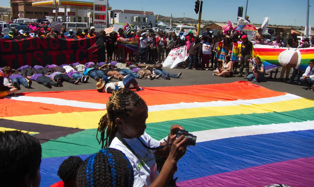 A very large flag spread flat across a road, six rainbow bands crossed by a white pall with a black wedge, with marchers lying face down on the tarmac beyond it beneath a banner reading DYING FOR JUSTICE.