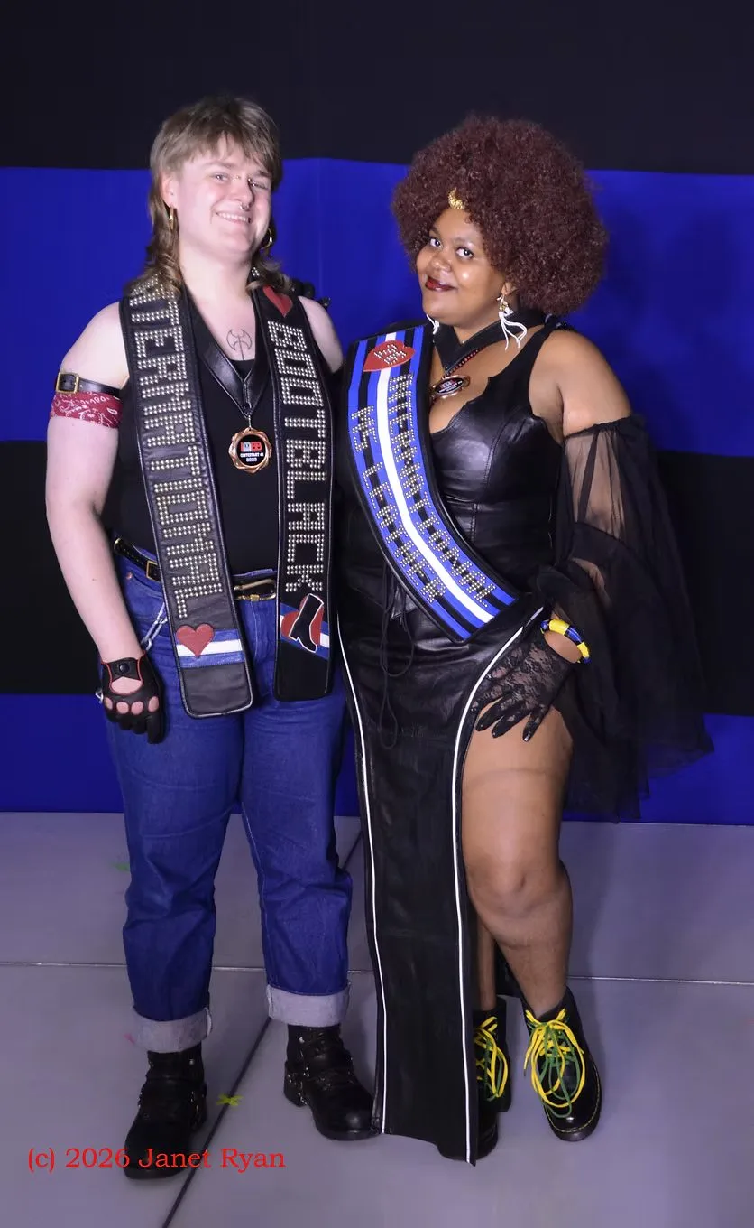 Two titleholders pose before a wall-sized Leather Pride Flag backdrop of black and blue stripes. At the left, a person in a black tank and rolled jeans wears a studded sash lettered INTERNATIONAL BOOTBLACK with a red heart and a bootblack patch, and a contestant medal. At the right, a person in a black leather dress with a high side slit and lace gloves wears a sash striped as the Leather Pride Flag lettered INTERNATIONAL MS LEATHER with a red heart pin. A watermark at the lower left reads (c) 2026 Janet Ryan.