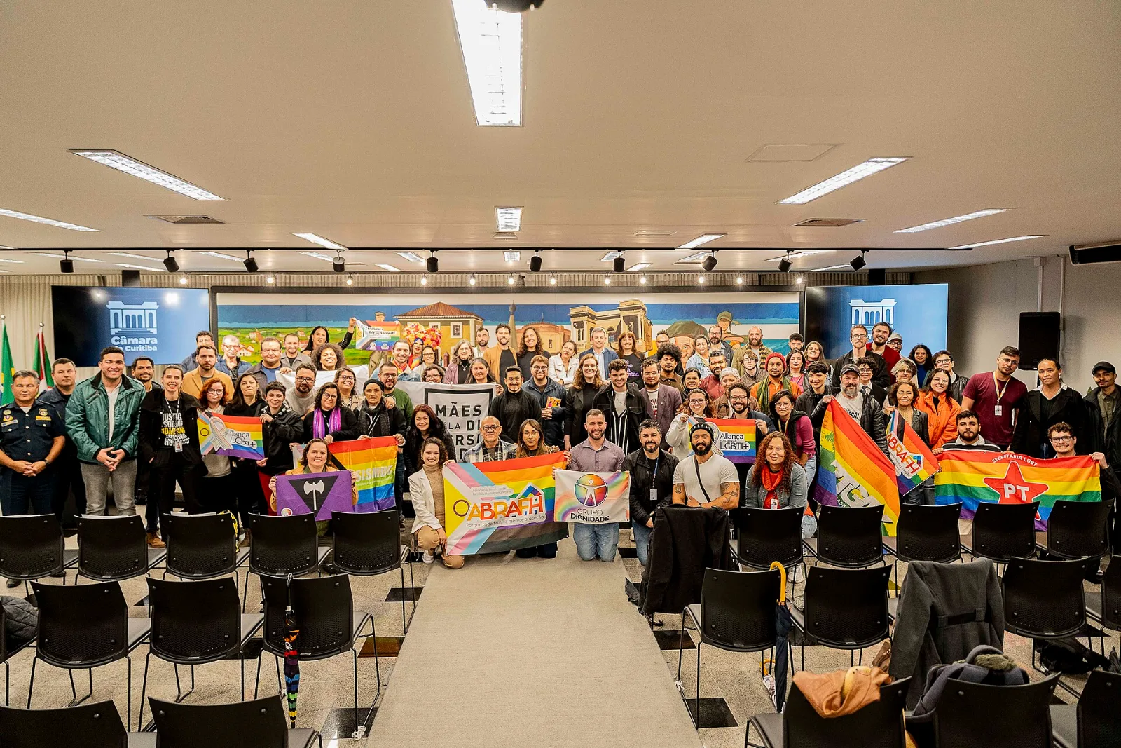 A large group photograph in a city council chamber, rows of black seats in the foreground. Toward the left of the group two people hold a violet flag with a black inverted triangle and white double-headed axe. Around it are rainbow banners lettered ABRAFH, Grupo Dignidade, Aliança LGBTI+ and Secretaria LGBT PT, and a black banner reading MÃES DA DIVERSIDADE.