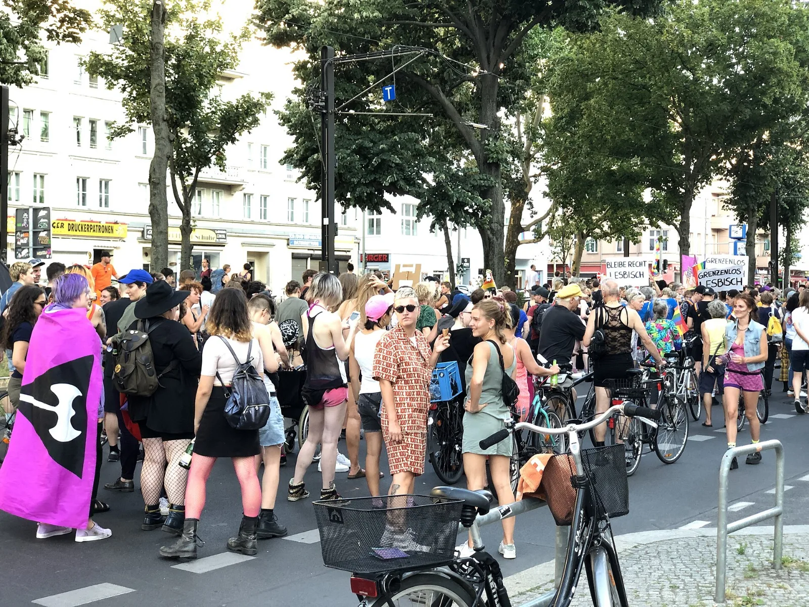 A person at the head of a crowded street march wears a magenta flag as a cape, its black inverted triangle and white double-headed axe turned toward the camera. Marchers with bicycles fill the road behind; hand-lettered placards at the right read BLEIBE RECHT LESBISCH and GRENZENLOS LESBISCH.