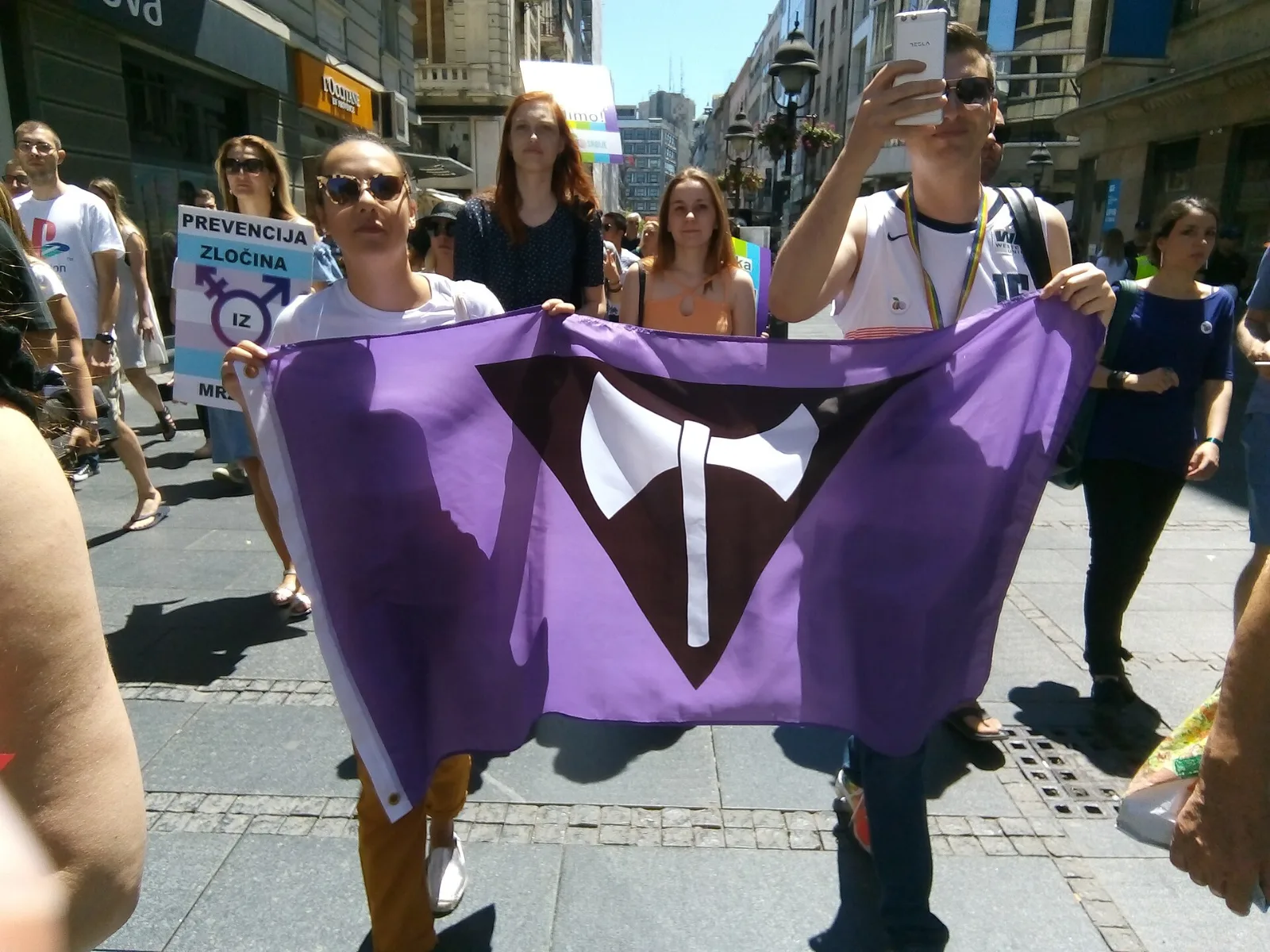 Two people carry a violet flag stretched between them along a sunlit city street, its black inverted triangle and white double-headed axe facing the camera. A crowd walks behind them; at the left a placard in pale blue, pink and white stripes reads PREVENCIJA ZLOČINA IZ MRŽNJE beside a combined gender symbol, and at the right a man films the march on a phone.