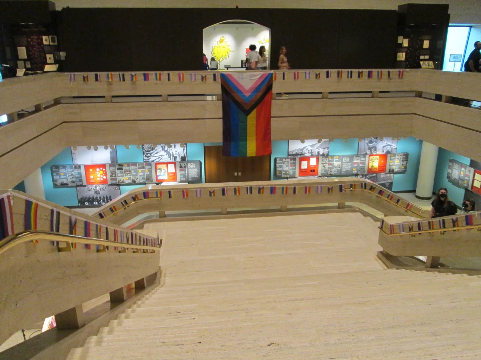 A row of pride flags hanging vertically from a high ceiling in a museum atrium, the transgender flag among them.