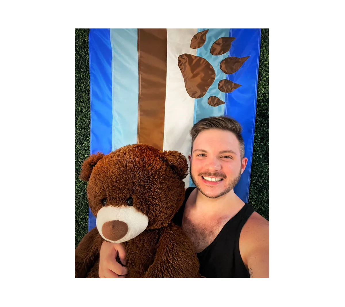 A man in a black tank top holds a large brown teddy bear in front of a sewn otter pride flag hung on a hedge wall, its stripes and paw print made of appliquéd panels.