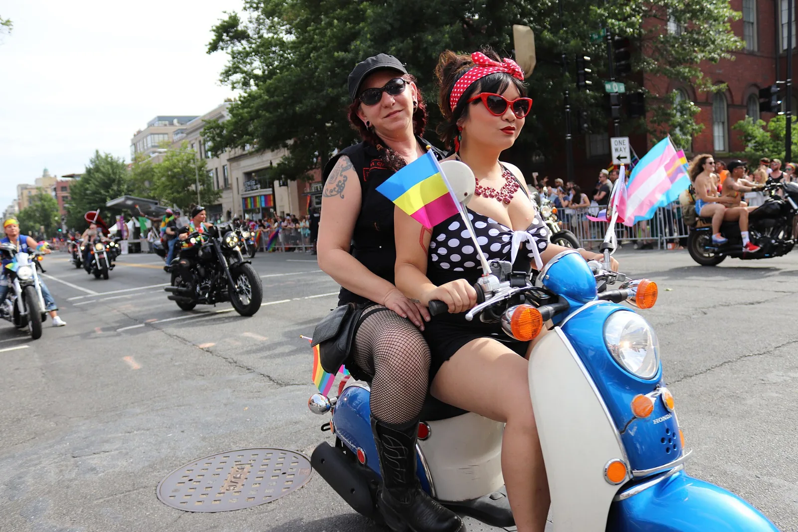 Two riders on a blue scooter at a Pride parade. On the handlebar mast a pansexual flag flown with light blue at the top and pink at the bottom, beside a transgender flag; a rainbow flag lower on the frame.