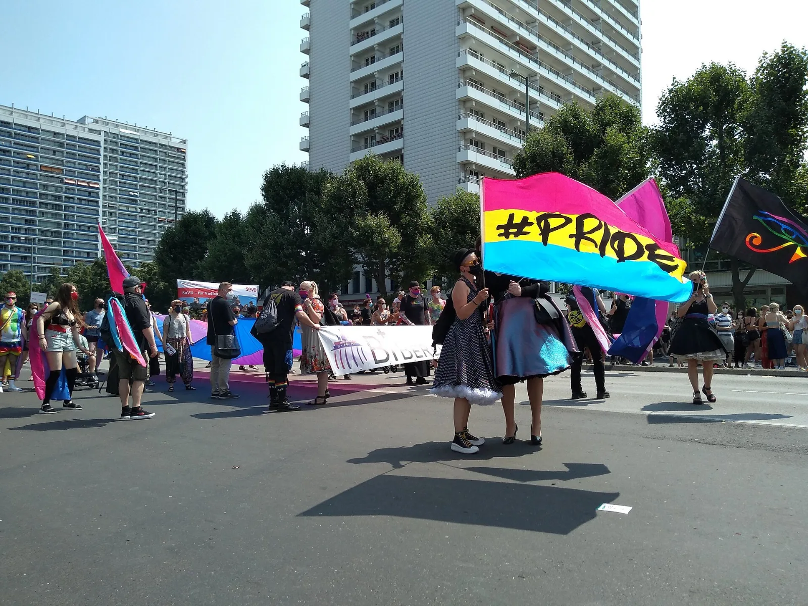 Two people carrying a large pansexual flag hand-lettered with the word PRIDE in black across the yellow and blue stripes, with bisexual flags and a Bi Berlin banner behind them.