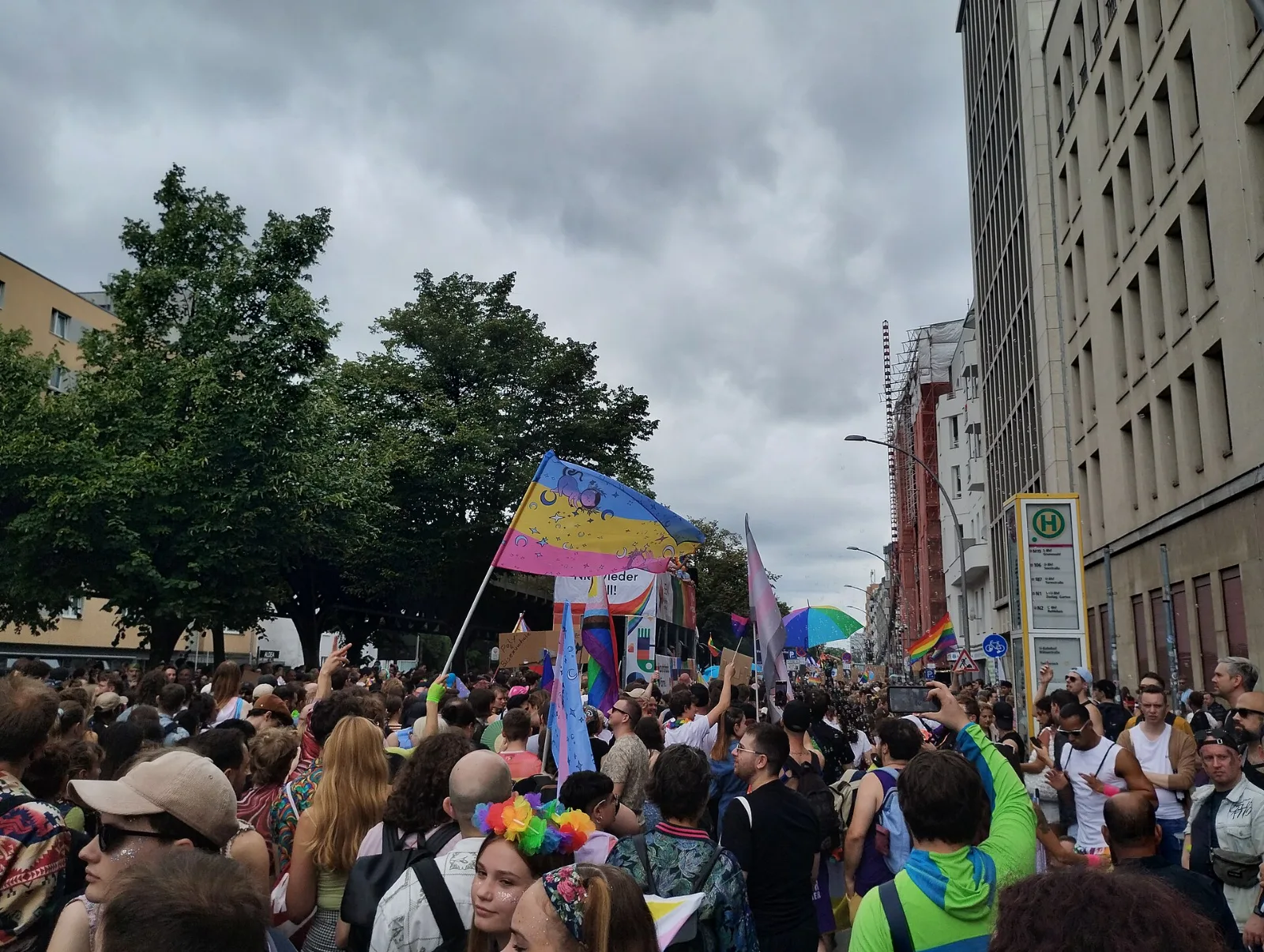 A large pansexual flag above a dense crowd, flown with light blue at the top and hand-painted with moons, stars and figures across all three stripes.