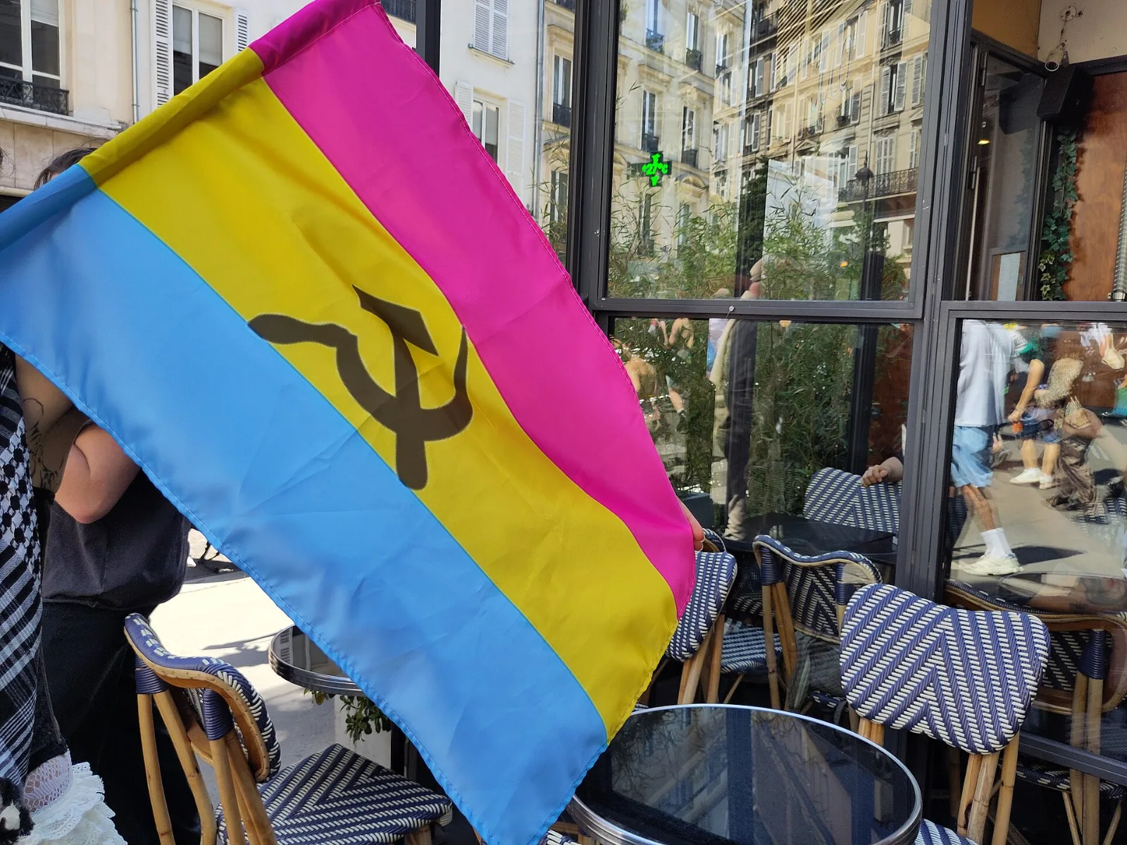 A pansexual flag held up outside a café, its yellow stripe charged with a dark hammer and sickle.
