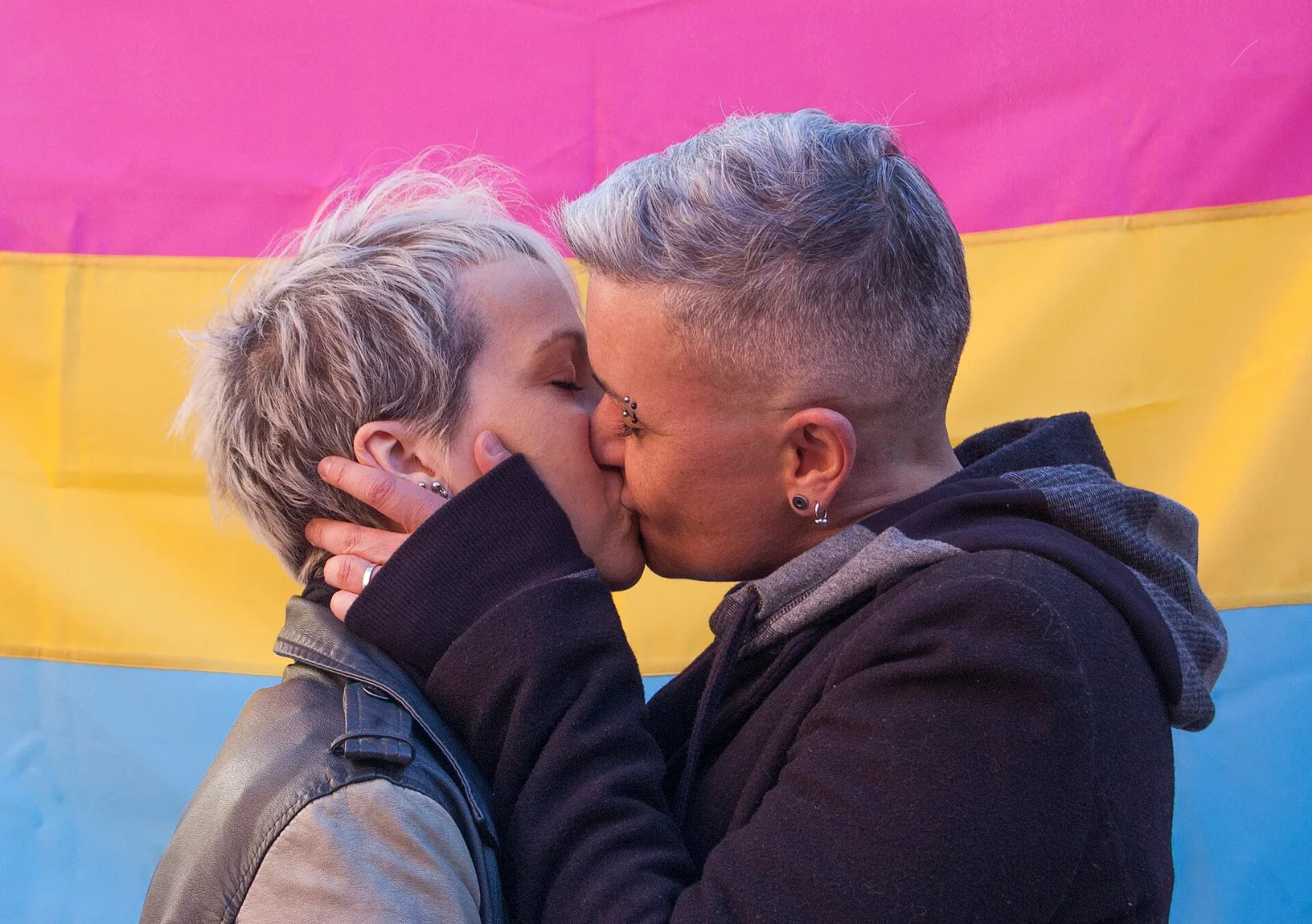 Two people with short pale hair kissing in front of a large pansexual flag hung as a backdrop, pink above yellow above light blue.