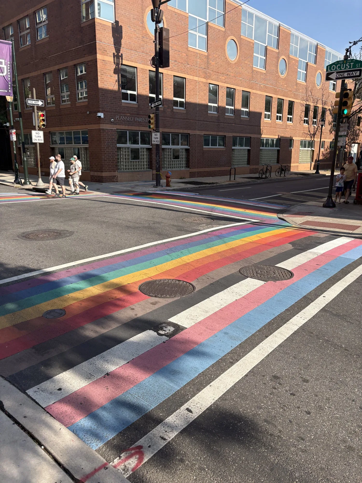 The Philadelphia Pride Flag crosswalk mural at 12th/13th and Locust Streets in Philadelphia's Gayborhood, photographed in daylight with the black and brown stripes leading into the traditional rainbow