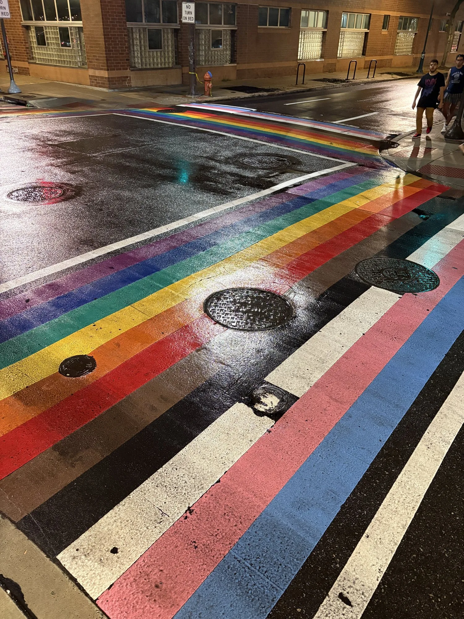 The Philadelphia Pride Flag crosswalk mural at 12th/13th and Locust Streets in Philadelphia's Gayborhood at night, its stripes reflecting on wet pavement under streetlight