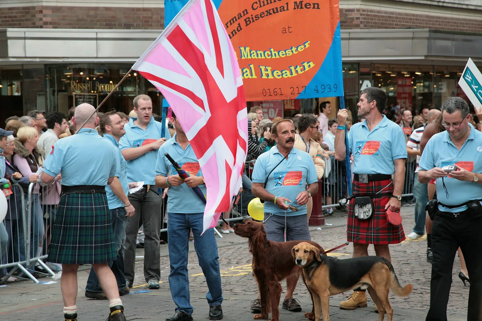 Marchers in kilts and light blue polo shirts at Manchester Pride carrying a large Pink Jack behind a sexual-health banner.