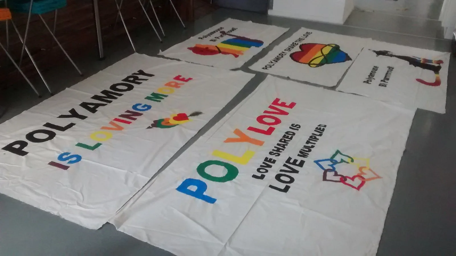 Hand-painted cloth banners laid out on a floor reading POLYAMORY IS LOVING MORE and POLY LOVE, several with infinity hearts.