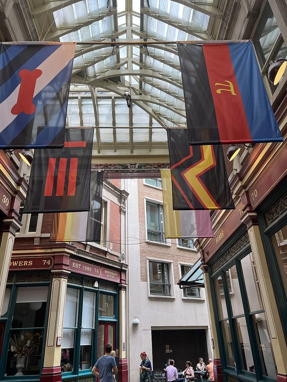 Large printed banners hanging beneath the glass roof of a Victorian market arcade, one of them blue, red and black with a stylised gold pi.