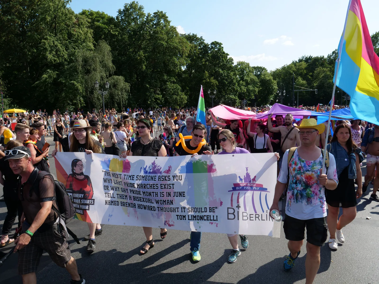 Marchers behind a white banner whose logo reads non-mono bi+ pan ply above the name BiBerlin. A pansexual flag is carried at the right and a partly furled pink, green and blue flag rises at the centre distance.