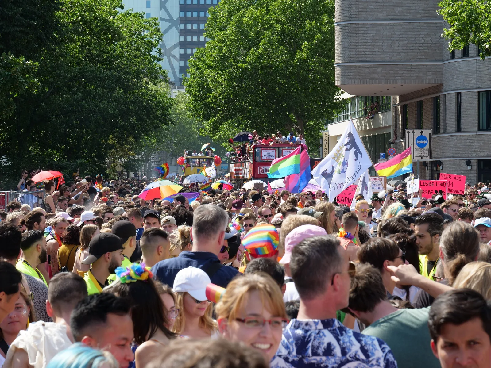 A crowded street parade. Above the crowd a three-stripe flag in pink, green and blue flies beside a pink, purple and blue bisexual flag, with a pansexual flag at the right.