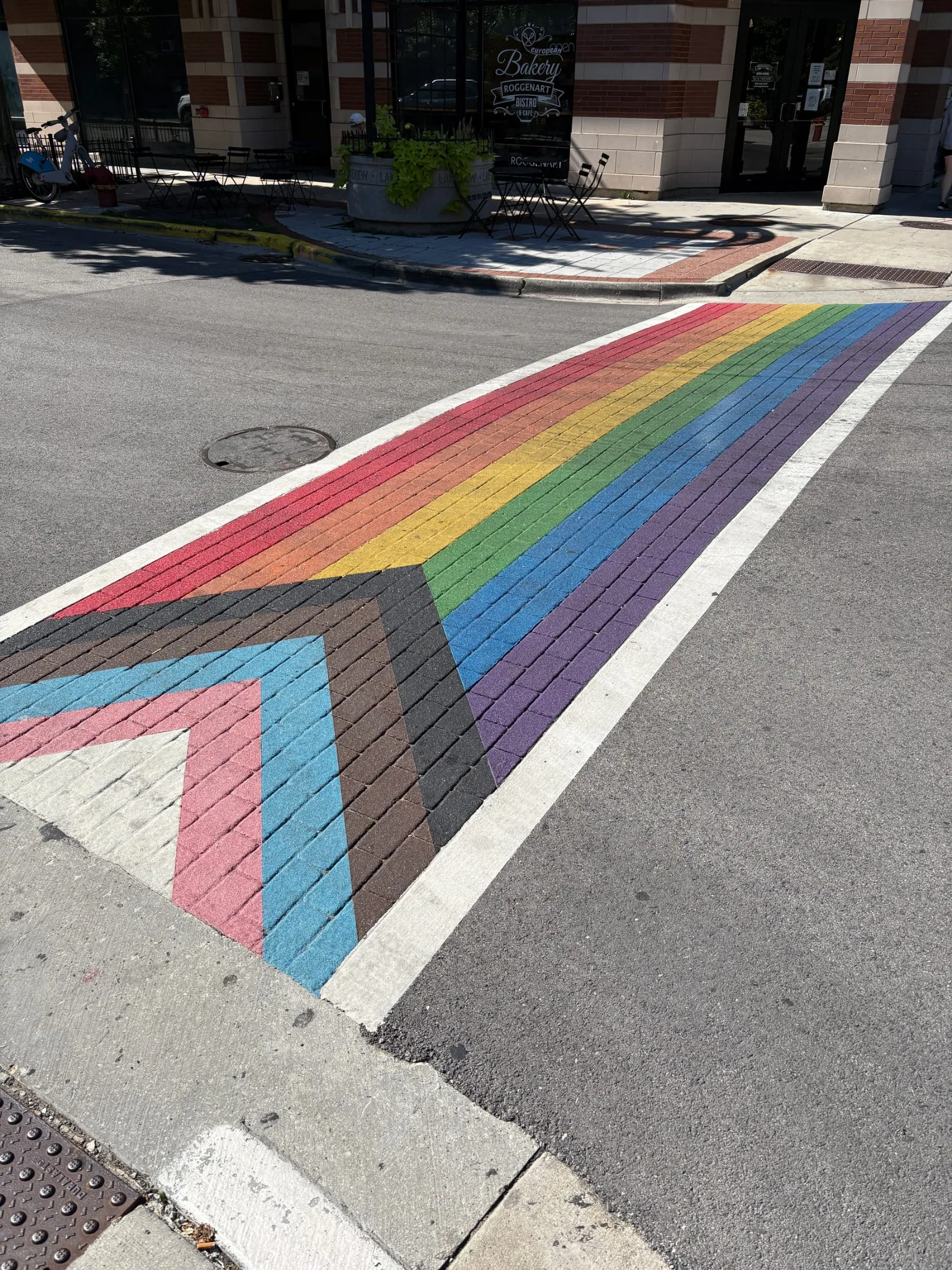 A Progress Pride crosswalk on North Halsted Street in Chicago: six rainbow stripes crossed by a chevron of white, pink, light blue, brown and black pointing in from the near corner, laid into brick paving outside a bakery.
