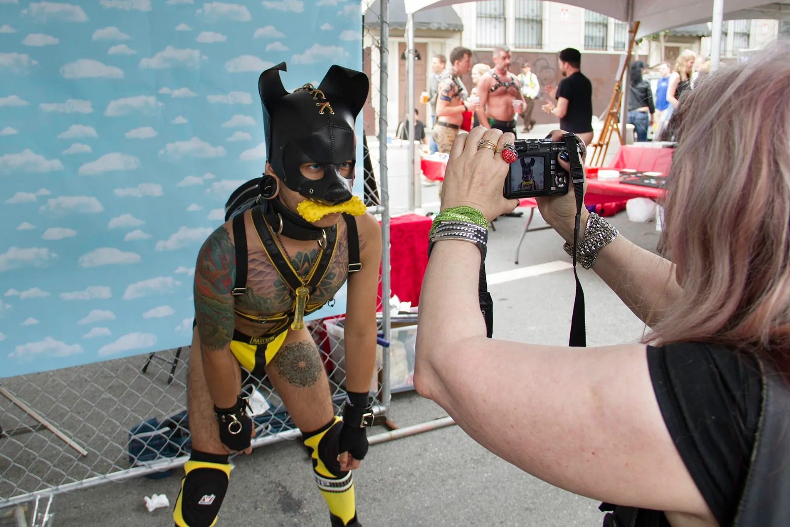 A person in a black pup hood and yellow harness being photographed at an outdoor street fair.